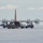 A large four-engine turboprop aircraft, consistent with a C-130 Hercules variant, is parked on a snow and ice surface in what appears to be a polar or Antarctic environment. Ground support vehicles and equipment surround the aircraft, which has its cargo ramp open, indicating active loading or unloading operations. The aircraft features an orange and red tail livery, suggesting affiliation with a polar research or support program.