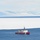 A U.S. Coast Guard vessel, painted in the distinctive red and white icebreaker livery, navigates open blue water at the edge of a large ice shelf or sea ice formation. Snow-covered mountains are visible in the background under a pale blue sky. The scene strongly suggests a polar or sub-polar environment, likely the Arctic or Antarctic region.