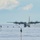 A large four-engine turboprop aircraft, consistent with a C-130 Hercules configuration, is taxiing or landing on a snow and ice runway in what appears to be a polar or arctic environment. The runway is marked with green and black flags/markers on both sides to delineate the landing strip on the featureless white terrain. All four propellers are visibly spinning, and snow/ice dust is being kicked up behind the aircraft.