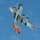 A bottom-up view of a white and orange US Air Force multi-engine turboprop aircraft in flight against a partly cloudy blue sky. The aircraft is photographed from directly below, revealing its underside markings, four spinning propellers, and distinctive orange tail markings. The livery and configuration suggest this is likely a Lockheed C-130 Hercules or similar large military transport aircraft.