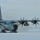 A U.S. Air Force LC-130 Hercules ski-equipped transport aircraft is shown on a snow and ice surface with all four turboprop engines running, as evidenced by the motion-blurred propellers. The aircraft bears the serial number 3903 and features the distinctive ski-wheel combination landing gear visible beneath the fuselage. Snow is being blown across the surface by propeller wash, suggesting the aircraft is preparing for takeoff or has recently landed in a polar environment.
