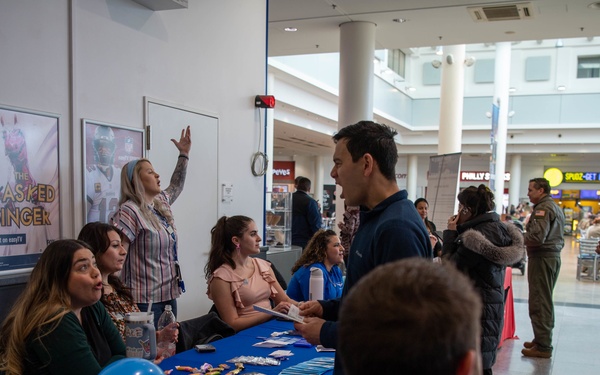 A busy indoor event or fair taking place in what appears to be a shopping mall or airport terminal. Multiple booths with blue tablecloths are staffed by participants distributing materials and engaging with visitors. Blue balloons, promotional items, and printed materials are visible on the tables.