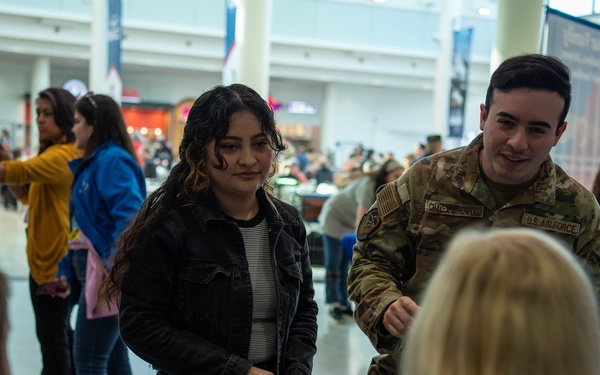 A U.S. Air Force service member in OCP (Operational Camouflage Pattern) uniform participates in what appears to be a military recruiting or community outreach event inside a shopping mall. A young woman in a black jacket is interacting with representatives at an information table covered with brochures and small items. A Subway restaurant is visible in the background, confirming the mall setting.