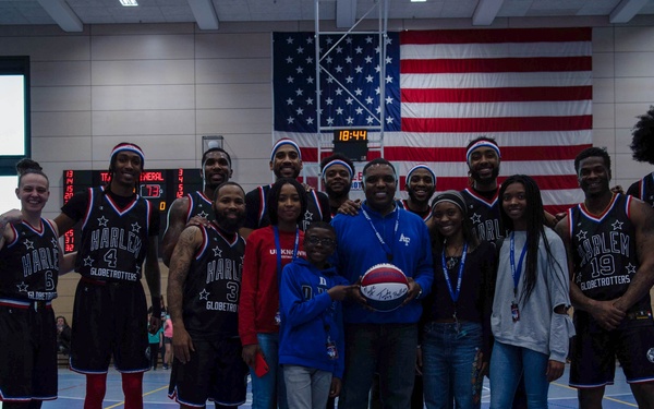 A group photo featuring Harlem Globetrotters players in their distinctive black, red, and white uniforms posing with what appear to be military personnel and civilians on a basketball court. A large American flag is prominently displayed on the wall behind the group, suggesting this is likely a military base visit or special event. One civilian in the center is holding a basketball, possibly a signed souvenir.