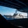 USS Carl Vinson (CVN 70) Fueling-at-sea The image is taken from inside a large naval vessel, likely an aircraft carrier, looking out through an open hangar bay or sponson opening at a U.S. Navy Arleigh Burke-class guided-missile destroyer sailing alongside. The destroyer displays hull number markings and has personnel visible on its deck, suggesting a replenishment-at-sea or close-formation sailing operation. An American flag is visible flying from the destroyer's mast.
