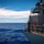 USS Carl Vinson (CVN 70) Sailor Stands Watch A naval vessel is underway on open ocean under clear blue skies. A crew member stands on the ship's exterior deck/bridge wing, using binoculars to scan the horizon. The image captures the starboard side of a gray military ship with safety and navigational equipment visible.