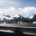 USS Carl Vinson (CVN 70) Flight Deck Operations The image shows a busy U.S. Navy aircraft carrier flight deck with multiple fixed-wing aircraft and a helicopter in operation. Deck crew members in colored jerseys are visible performing various tasks around the aircraft. The scene captures active flight deck operations under partly cloudy skies over open ocean.