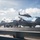 ARCHIVE -- USS Carl Vinson (CVN 70) Flight Deck Operations The image shows the flight deck of a U.S. Navy aircraft carrier with multiple fixed-wing aircraft and a helicopter in operation. Several deck crew members in colored jerseys are visible performing various flight deck duties around the aircraft. A helicopter, likely an MH-60 Seahawk, is airborne above the deck while jet aircraft, appearing to be F/A-18 Hornets or Super Hornets, are staged on the wet deck.