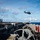 USS Carl Vinson (CVN 70) Flight Deck Operations A military helicopter, appearing to be an MH-60 Seahawk, is airborne above the flight deck of a naval vessel at sea. The image is taken from a lower deck perspective, showing deck equipment and machinery in the foreground with the flight deck elevated above. The ocean and partly cloudy sky are visible in the background.