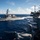 USS Carl Vinson (CVN 70) Fueling-at-sea The image shows a U.S. Navy Arleigh Burke-class guided-missile destroyer underway at sea, photographed from the deck of what appears to be a U.S. Navy aircraft carrier. Multiple naval vessels are visible, with aircraft parked on the carrier's flight deck in the foreground. The scene depicts a carrier strike group operating in open ocean under partly cloudy skies.