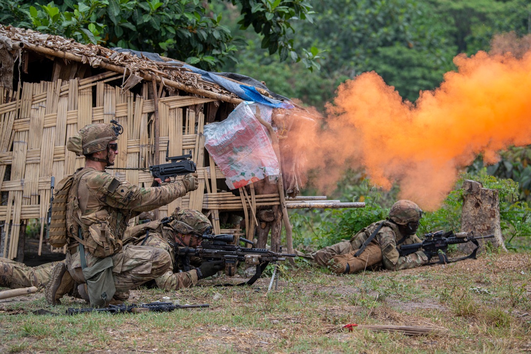 Soldiers with weapons kneel and lie on the ground as orange smoke floats through the air.