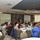 DPAA hosts Chicago FMU A formal conference or seminar is taking place in a modern banquet/meeting room with numerous attendees seated at round tables. A presenter stands at a podium at the front of the room, flanked by two projection screens displaying presentation slides and an American flag. The room features contemporary circular pendant lighting and large windows along the walls.