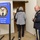 DPAA hosts Chicago FMU The image shows the entrance to a Defense POW/MIA Accounting Agency (DPAA) Korean War and Cold War Meeting. A U.S. Army officer in dress uniform stands near the doorway greeting attendees as two civilians enter the meeting room. A blue DPAA sign on a stand prominently displays the agency seal and meeting title.