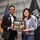 DPAA hosts Chicago FMU Two women are posing together while jointly holding a commemorative plaque or photo collage from the Defense POW/MIA Accounting Agency (DPAA). The setting appears to be a formal recognition or award ceremony, with an American flag and a POW-MIA flag visible in the background. The plaque appears to reference an April 22, 2023 Chicago/Illinois Family Member Update event.