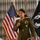 DPAA hosts Chicago FMU A U.S. Marine Corps officer in dress green uniform is speaking at a formal event while holding a microphone. He is flanked by an American flag with gold fringe on the left and a POW/MIA flag on the right. The setting appears to be an indoor ceremonial or commemorative event.