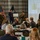 DPAA hosts Chicago FMU A formal military presentation or briefing is taking place in a conference or banquet hall setting. Two military personnel in Marine Corps dress uniforms and one Air Force officer at a podium are presenting to a seated civilian audience. A projection screen displays slides with text and images in the background.