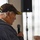 DPAA hosts Chicago FMU An elderly man with white hair is speaking into a handheld microphone at what appears to be a public event or gathering. He is wearing a dark brown baseball cap with yellow embroidery that reads 'United States Army' and a patterned blue sweater. The photo is taken from a rear three-quarter angle, with a blurred background showing a speaker stand and curtains.