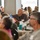 DPAA hosts Chicago FMU A large indoor gathering of predominantly older adults seated at round tables, likely a formal luncheon or commemorative event. A small American flag is visible on one of the tables, and several attendees are wearing name badges and veteran-style caps, suggesting a patriotic or veterans-related event. The attendees appear to be attentively listening to a speaker or presentation off-camera.