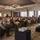 DPAA hosts Chicago FMU A large conference or seminar event is taking place in a modern hotel ballroom. A speaker stands at a podium on a raised stage addressing a full audience seated at round tables. The room is well-lit with distinctive circular ring pendant lights overhead.