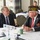 DPAA hosts Chicago FMU Two older men are engaged in conversation at a formal meeting or conference table labeled 'Table 1' with a 'Government Officials' placard visible. The man on the right is wearing a military veterans cap inscribed 'State Commander 2022-2023 Illinois,' indicating this is a veterans organization event. The setting appears to be a hotel or conference center banquet room with additional attendees visible in the background.