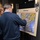 DPAA hosts Chicago FMU Two individuals are examining a display board titled 'POW Camps' showing a map of prisoner of war camp locations, likely related to the Korean War. The map includes labeled camp locations such as 'Air Pirate Prison,' 'Manpo Camps,' 'Apex Camps,' 'Kanggye,' 'Valley #1,' and 'Suan Camps.' The display appears to be part of a formal briefing or memorial event, featuring official insignia including what appears to be a DoD seal and a POW/MIA emblem.
