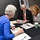 DPAA hosts Chicago FMU Three professionals are seated around a dark table engaged in what appears to be a formal meeting or review session. Documents, folders, and printed materials are spread across the table, suggesting a structured discussion or evaluation. A banner in the background references a 'Medical Examiner System' and 'Delaware,' indicating an official or governmental context.