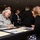 DPAA hosts Chicago FMU An indoor gathering or event, likely a registration table or information booth, where a woman in a gray hoodie is seated at a black-draped table writing on a form or document. A younger woman with long auburn hair leans over the table, appearing to assist or interact with her. The background shows a crowded room with multiple attendees milling about.