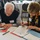 DPAA hosts Chicago FMU Two elderly individuals are seated at a round table covered with documents, forms, and folders, actively filling out paperwork. Both wear name badges and appear to be at an organized event or registration session. The setting appears to be a banquet hall or conference room with additional tables visible in the background.