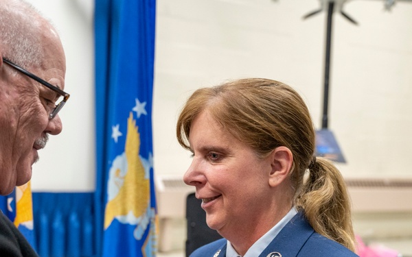 A civilian man is pinning a medal or insignia onto the uniform of a female U.S. Air Force senior enlisted member during what appears to be a formal military ceremony. The woman is wearing a dress blue uniform with multiple medals and ribbons, and her rank insignia on the sleeve indicates she is a Chief Master Sergeant. The setting includes a blue Air Force flag in the background, suggesting an official indoor ceremony venue.