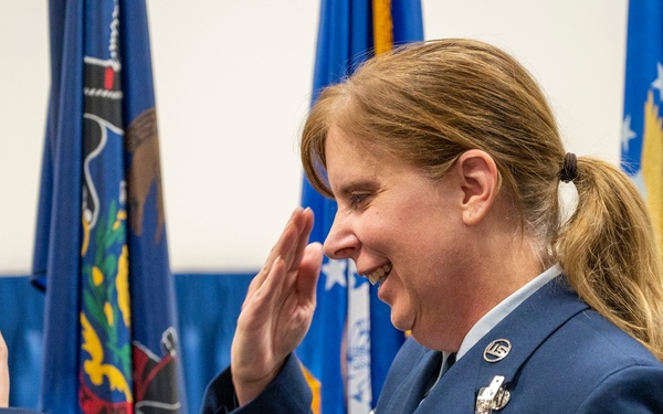 Two female U.S. Air Force personnel are exchanging salutes during what appears to be a formal military ceremony. The individual on the right, wearing the rank of Chief Master Sergeant (E-9), is smiling and displaying multiple service ribbons and medals. Blue military flags, including what appears to be a Pennsylvania state flag and Air Force flags, are visible in the background.