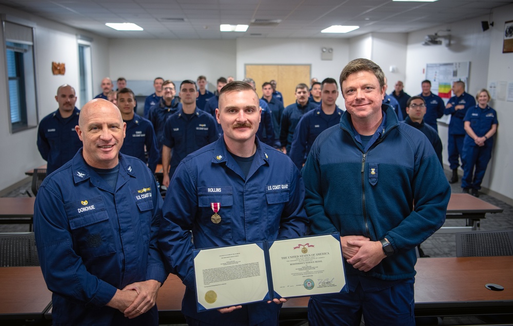 Coast Guard Capt. Keith Donohue, commander of Sector Houston-Galveston, Petty Officer 2nd Class Chad Rollins, a machinery technician at Station Sabine, and Master Chief Petty Officer Steven Beasley, officer-in-charge of Station Sabine, pose for a photo in front of the station crew during an award ceremony in Sabine Pass, Texas, Dec. 13, 2023. Rollins received a Meritorious Service Medal for rescuing two men from a sinking tugboat on March 31, 2023. (U.S. Coast Guard photo by Petty Officer 1st Class Corinne Zilnicki)