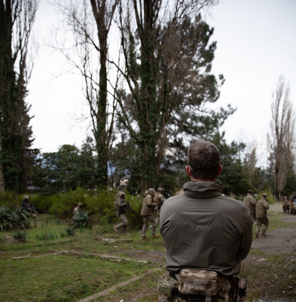 A U.S. MARSOC Marine Raider observes Georgian, and Spanish special operations forces soldiers move out to practice raiding a compound during Trojan Footprint 24