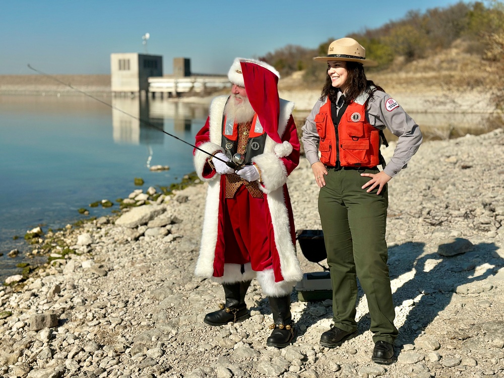 Santa Claus fishing while wearing a life jacket and standing next to USACE park ranger.