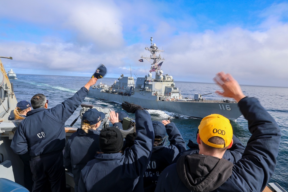 Cmdr. Arturo Trejo and and U.S. Navy Sailors wave to  the crew onboard the USS Thomas Hudner (DDG 116)
