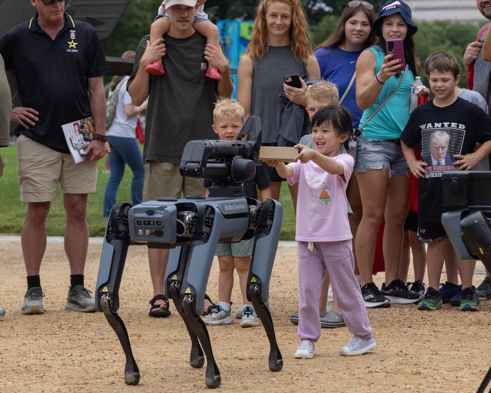 A little girl hands a stick -like object to a Boston Dynamics robotic dog at the 250th U.S. Army Birthday Festival