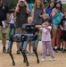 A little girl hands a stick -like object to a Boston Dynamics robotic dog at the 250th U.S. Army Birthday Festival