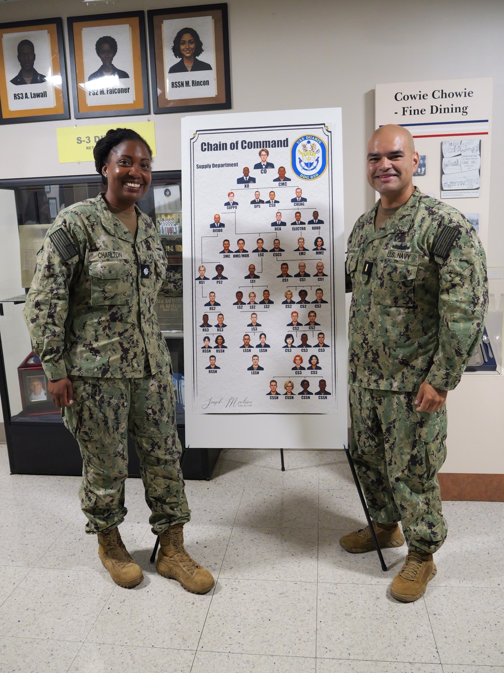 NSCS Executive Officer CDR Jennifer Charlton (left) and artist ENS Joseph Montano (right) stand next to an illustration containing the full crew of the USS Duarte (DDG 250)
