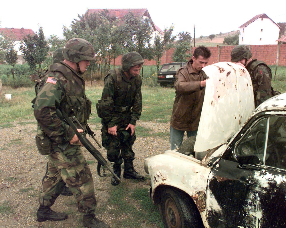 Soldiers surround a car while the owner opens the hood for inspection.