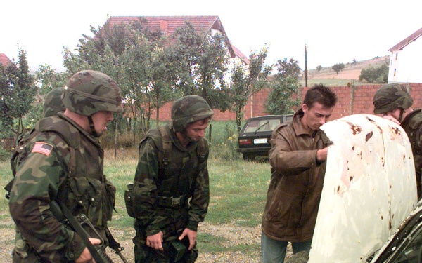 Soldiers surround a car while the owner opens the hood for inspection.
