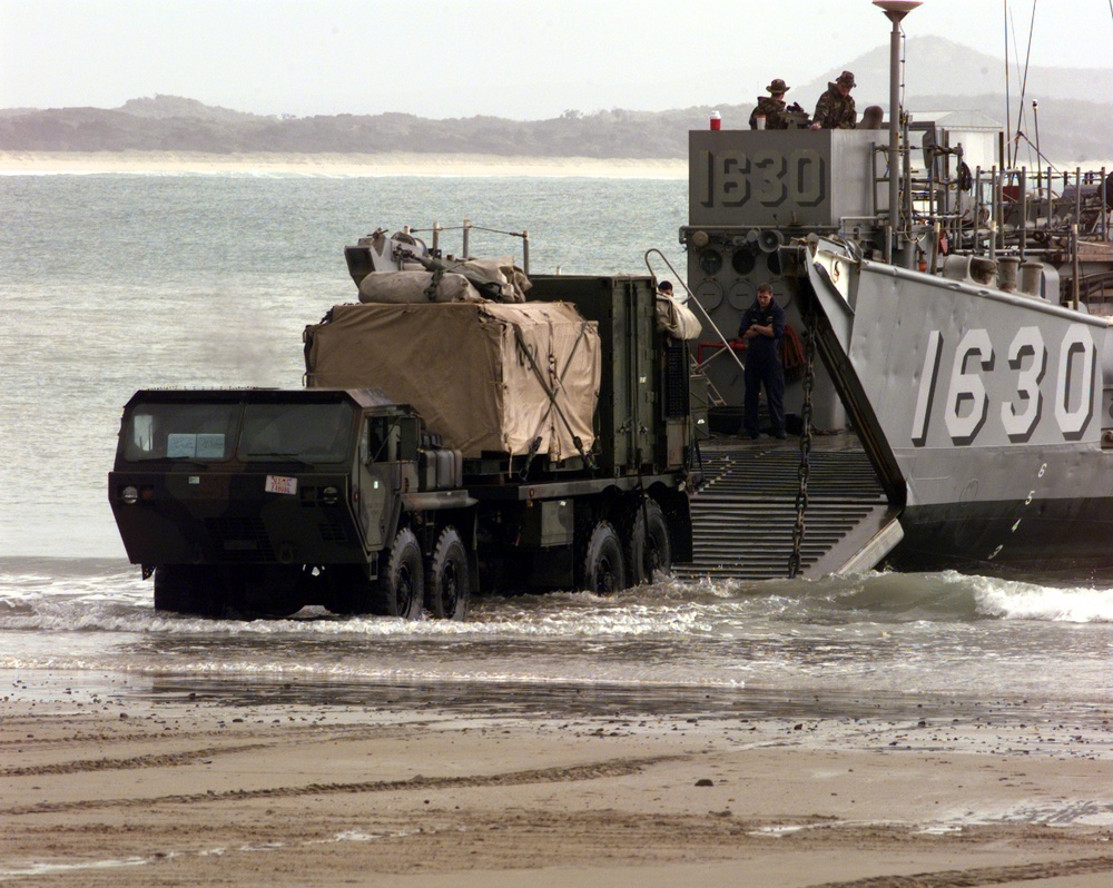 A U.S. Marine Corps logistics vehicle drives down the ramp of a U.S. Navy Landing Craft Utility into the surf at Freshwater Beach.