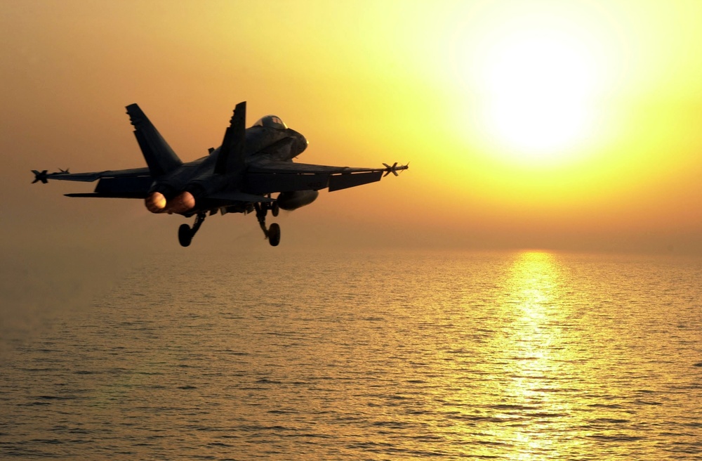 An F/A-18 Hornet launches towards the sun from the flight deck of the USS Constellation.