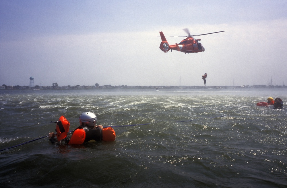 An HH-65A Dolphin helicopter lowers Petty Officer 3rd class Zee Lee to pick up aircrew members.