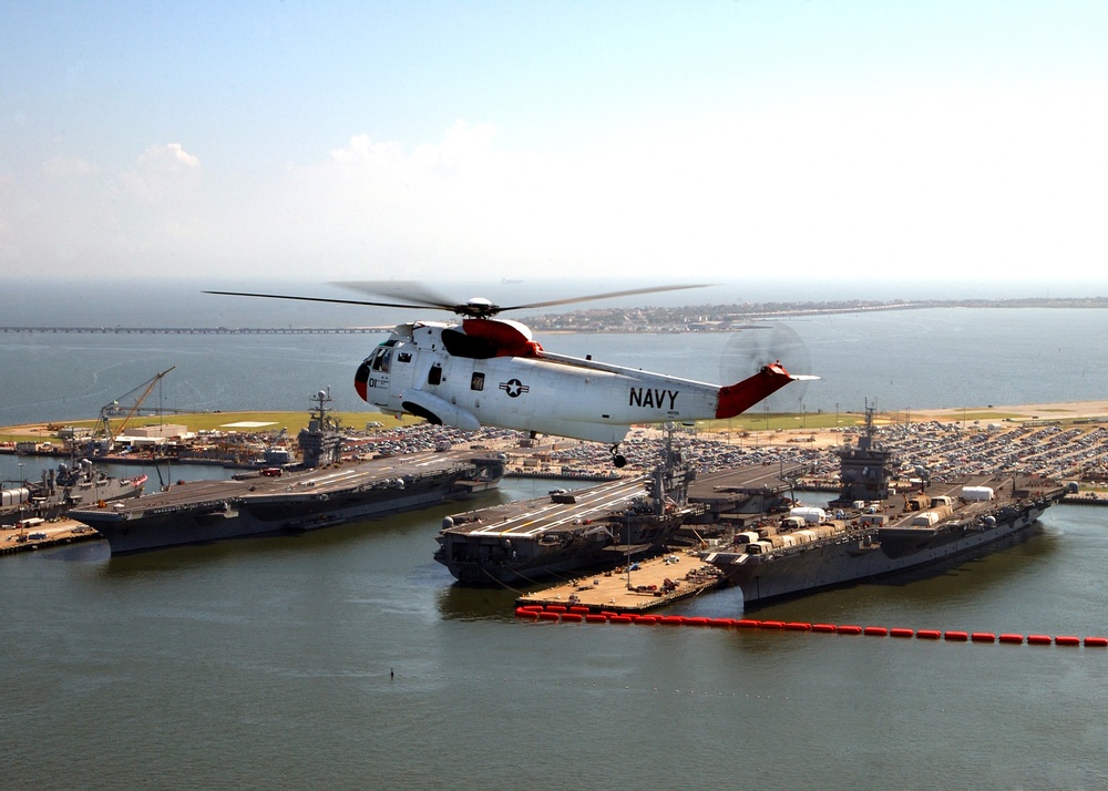 A UH-3H Sea King helicopter flies near three aircraft carriers moored at Naval Station Norfolk.