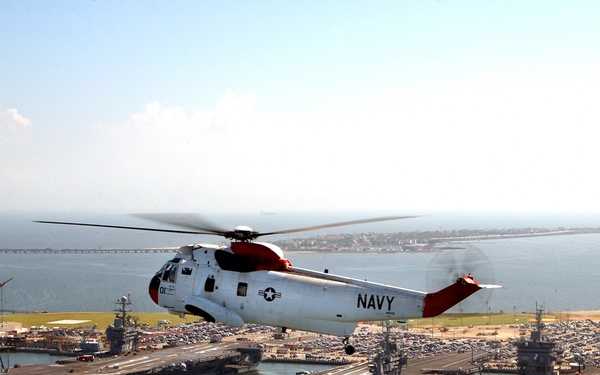 A UH-3H Sea King helicopter flies near three aircraft carriers moored at Naval Station Norfolk.