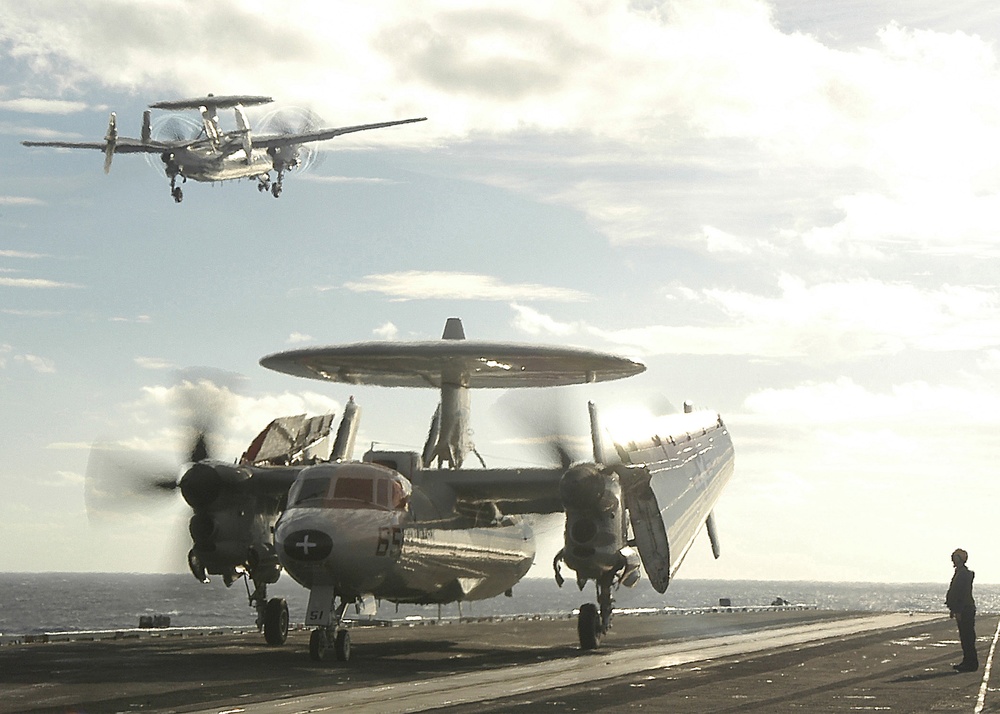 One Navy E-2C Hawkeye performs touch and go landings while another Hawkeye awaits taxi directions.