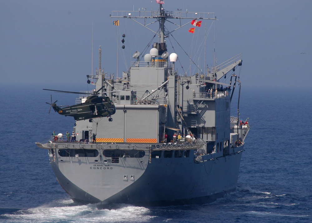 A crewman on the USNS Concord (T-AFS 5) hooks a cargo net of supplies to a UH-3H Sea King helicopter.