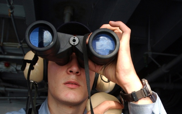 Navy Seaman Keith Ketterer uses his binoculars to search the horizon for any air or surface contacts.