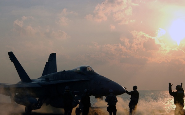 A U.S. Navy flight deck handler guides an F/A-18 Hornet aircraft into position for a catapult launch.