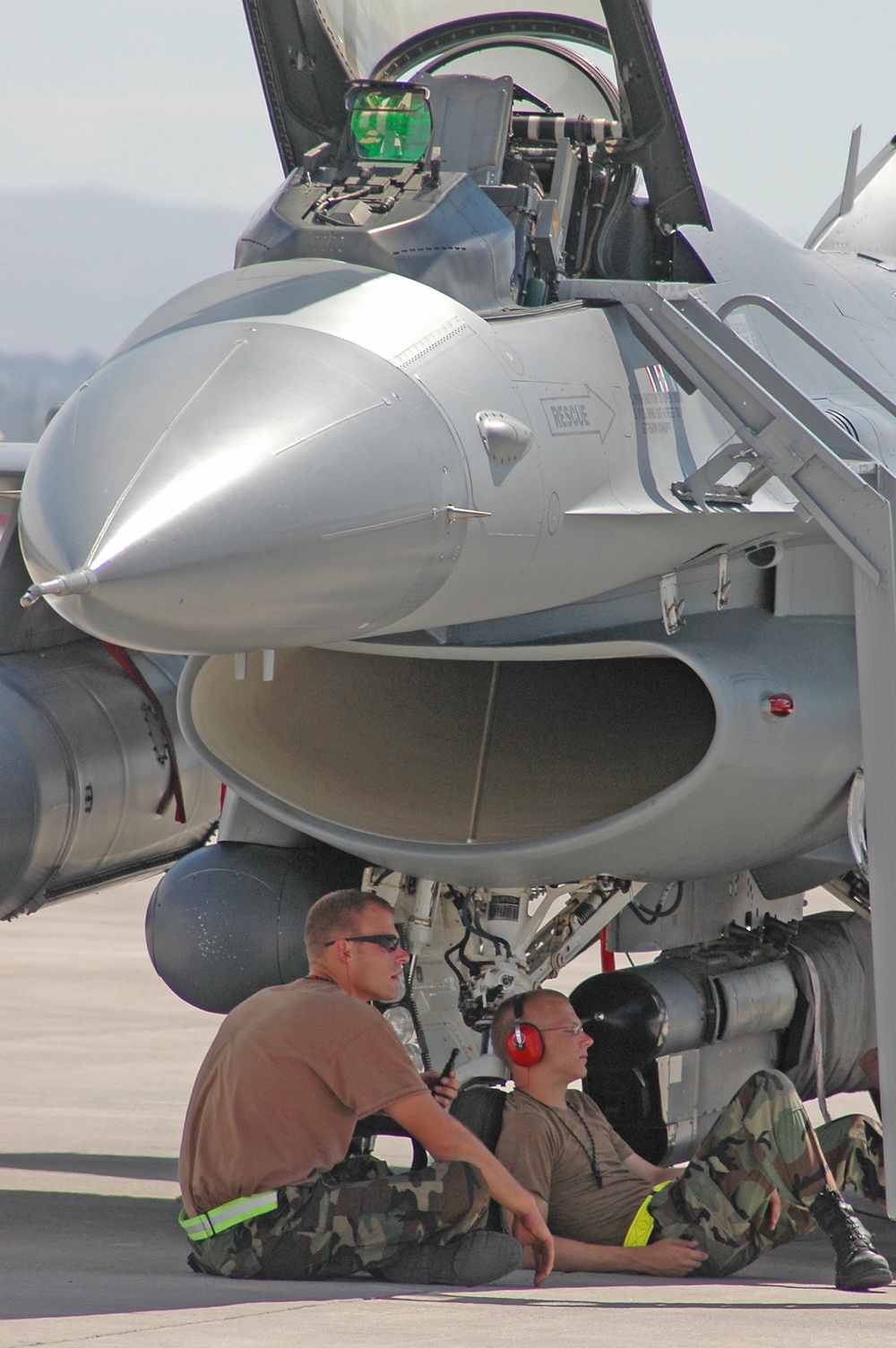 Senior Airmen Brent Bott and Zack McLaughlin seek shade under an F-16 Fighting Falcon.