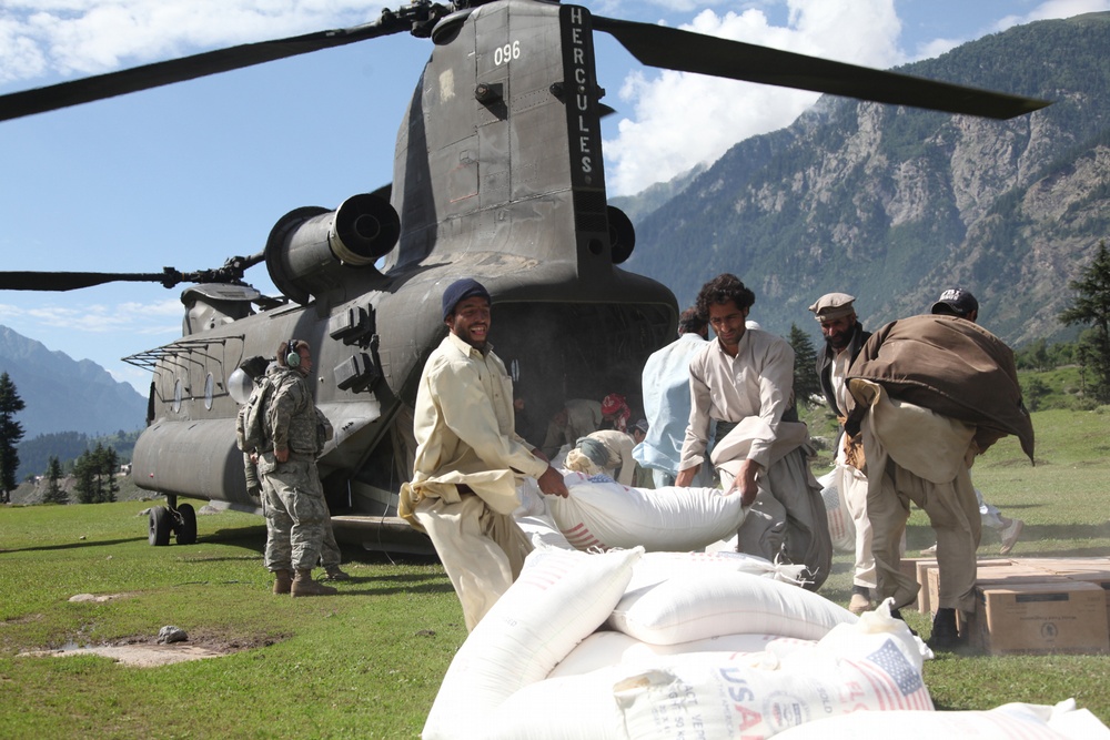 Pakistani men throw a bag of flour onto a pile behind a U.S. Army CH-47 Chinook helicopter.